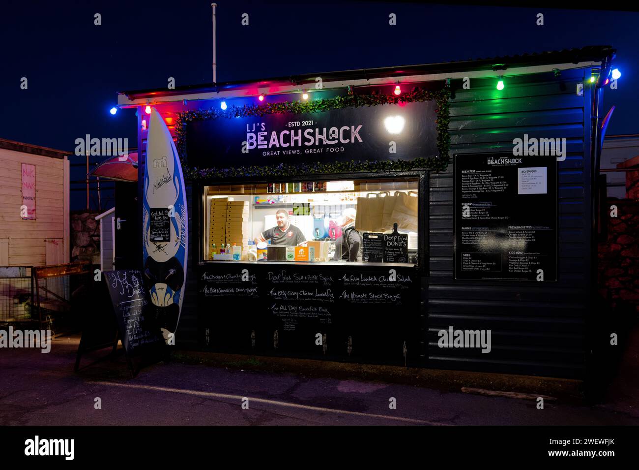 Le kiosque de restauration rapide Beach Shack à Teignmouth, Royaume-Uni la nuit. Un mâle et une femelle servent dans l'écoutille avec des menus à l'extérieur du kiosque. Banque D'Images