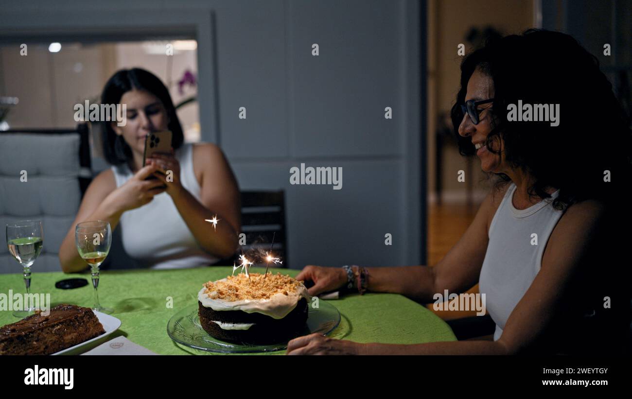 Deux femmes célèbrent à l'intérieur avec un gâteau aux bougies étincelantes, capturant un moment décontracté et joyeux dans un cadre confortable. Banque D'Images