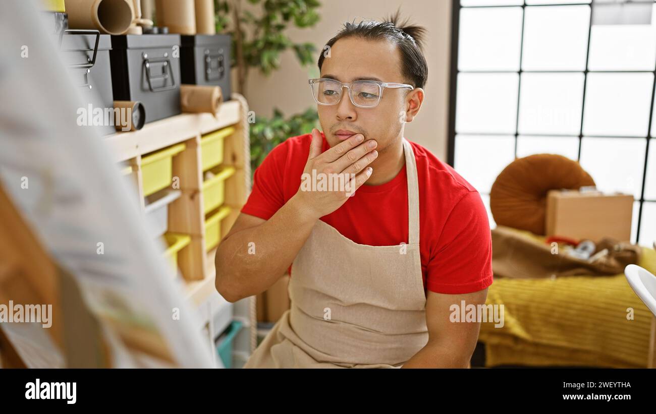 Pensées complexes d'un jeune artiste chinois, absorbé par l'art du dessin, habillé d'un tablier et de lunettes dans son studio confortable Banque D'Images