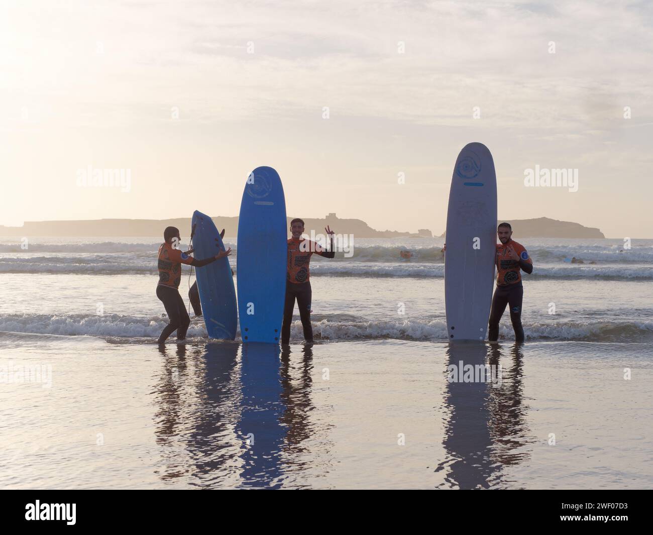 Trois apprenants surfeurs masculins sourient alors qu'ils tiennent leurs planches de surf comme pose pour une photo de plage au coucher du soleil à Essaouira, Maroc, le 27 janvier 2024 Banque D'Images