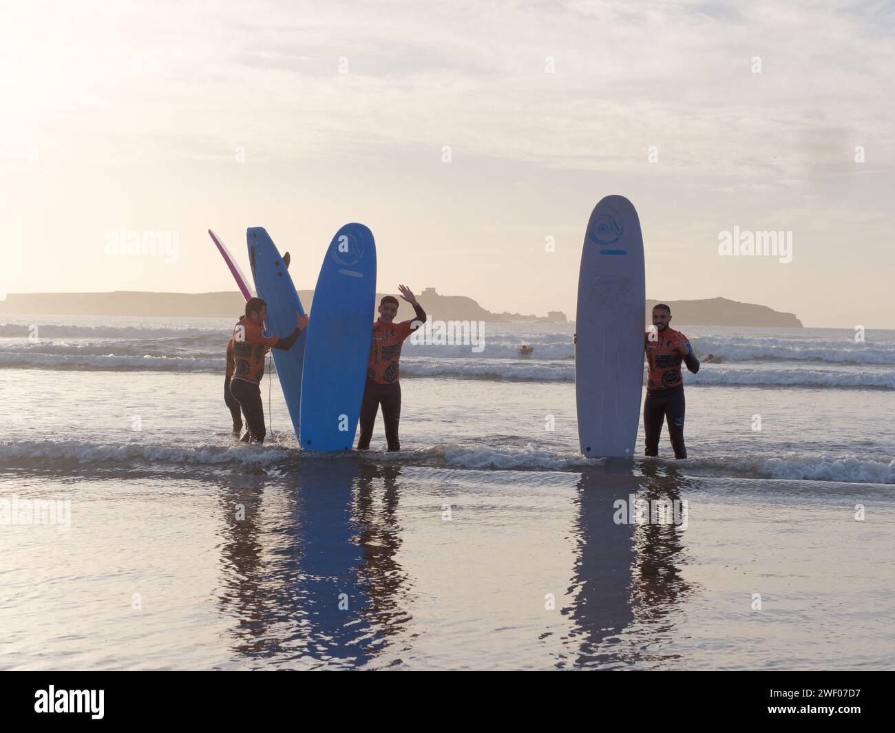 Trois apprenants surfeurs masculins sourient alors qu'ils tiennent leurs planches de surf comme pose pour une photo de plage au coucher du soleil à Essaouira, Maroc, le 27 janvier 2024 Banque D'Images