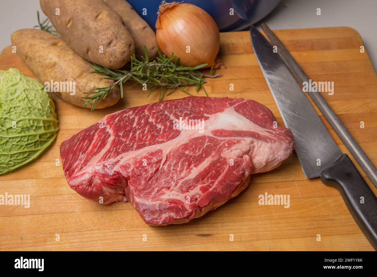 Rôti de bœuf sur une planche à découper en bois avec un couteau et de l'acier aiguisé. pommes de terre, chou, oignon et romarin. Banque D'Images