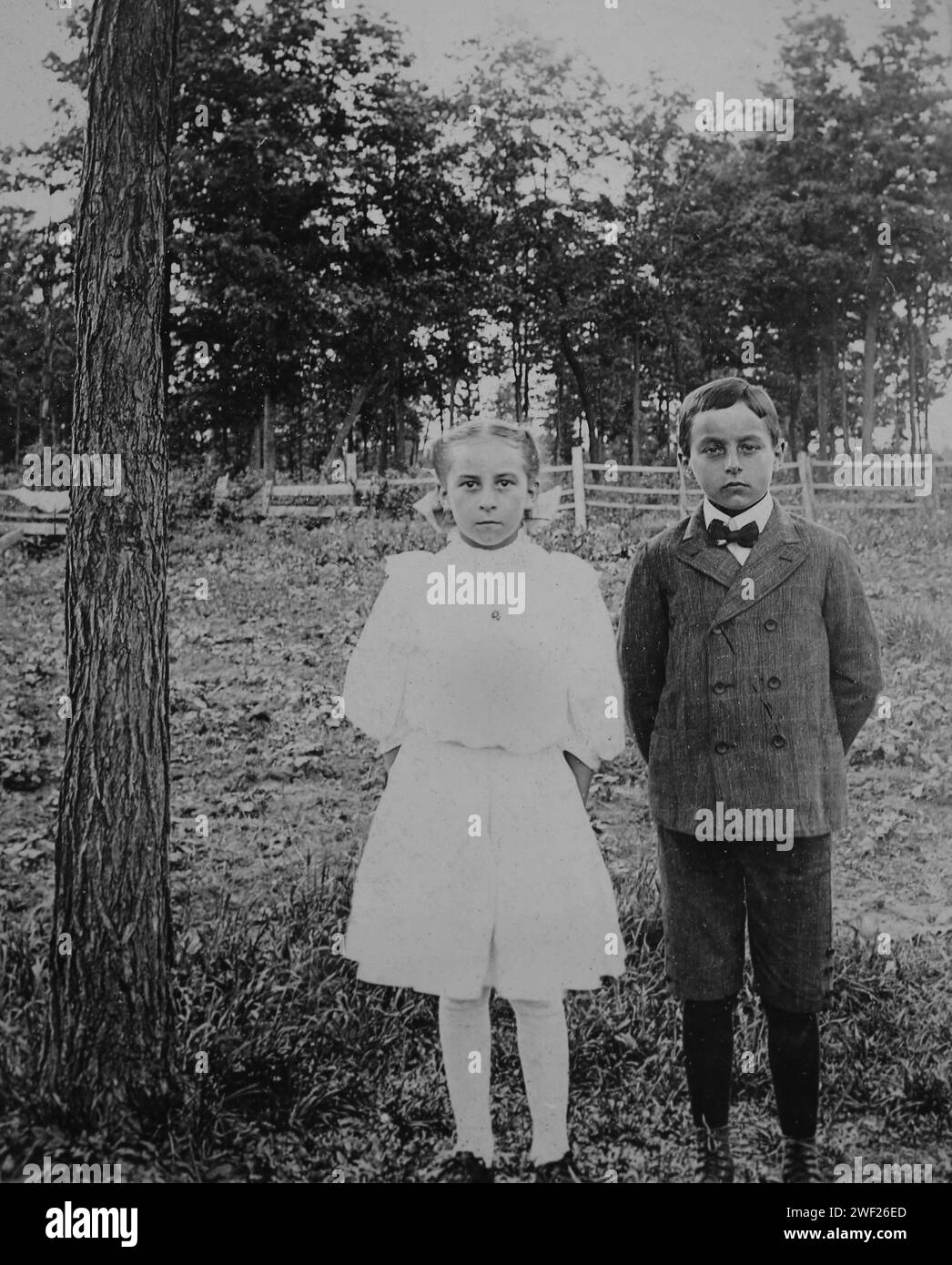 Deux enfants à l'apparence sévère posent debout juste à côté d'un arbre, CA. 1920. Banque D'Images