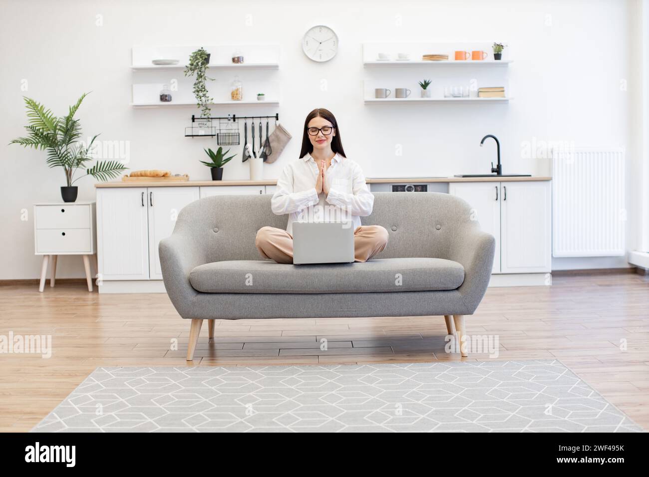 Brunette femme freelance assis sur un canapé confortable dans la pose de namaste avec la réduction de la fatigue en utilisant la pratique du yoga. Femme caucasienne méditant avec EY fermé Banque D'Images