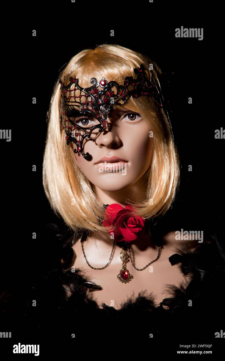 Mannequin femme en plastique avec les cheveux blonds brillants portant un demi-masque métallique noir avec des pierres rouges, et des vêtements romantiques et accessoires posant sur un bla Banque D'Images