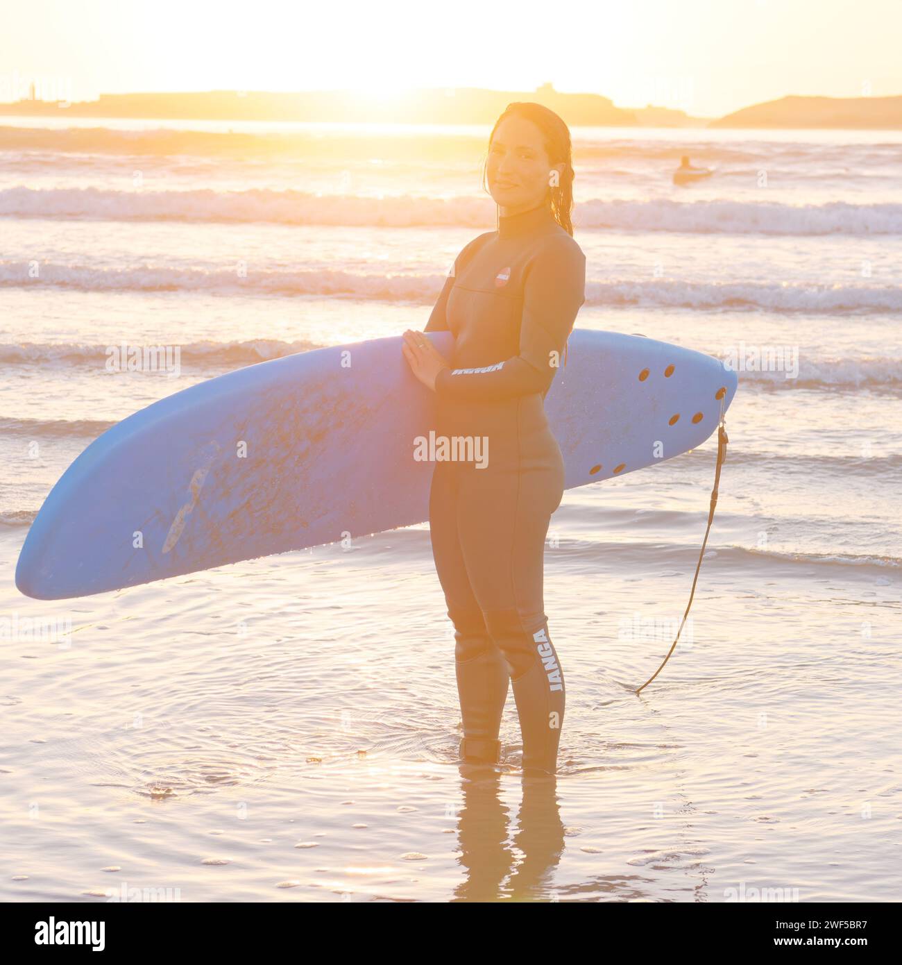 Surfeuse souriante en combinaison humide tient sa planche de surf au coucher du soleil à Essaouira, Maroc, le 28 janvier 2024 Banque D'Images