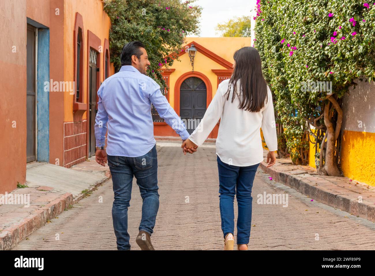 Vue arrière de couple marche main dans la main le long d'une rue colorée à Tequisquiapan Queretaro Mexique entouré de murs vibrants et de fleurs en fleurs Banque D'Images