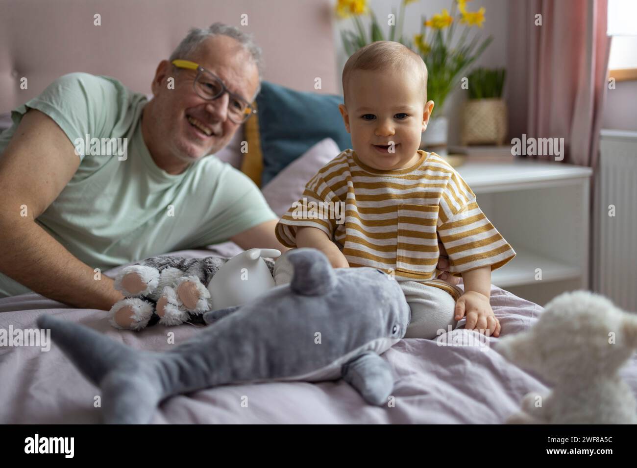 Un grand-père joyeux allongé à côté d'un enfant heureux, entouré de jouets en peluche dans une pièce confortable Banque D'Images