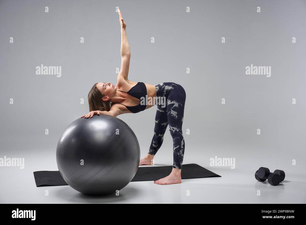 Jeune femme motivée dans le soutien-gorge de sport, en utilisant FIT ball pour l'entraînement. Vue latérale d'une fille sportive qui se penche, avec le bras levé, l'exercice, isolé sur un goujon gris Banque D'Images Jeune femme motivée dans le soutien-gorge de sport, en utilisant FIT ball pour l'entraînement. Vue latérale d'une fille sportive qui se penche, avec le bras levé, l'exercice, isolé sur un goujon gris Banque D'Images