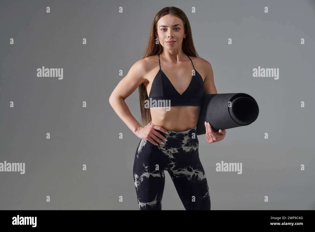Sportif souriant posant avec tapis de gymnastique à l'intérieur. Vue avant de l'athlète brune mince posant avec la main sur la taille, regardant l'appareil photo, pendant l'entraînement, iso Banque D'Images Sportif souriant posant avec tapis de gymnastique à l'intérieur. Vue avant de l'athlète brune mince posant avec la main sur la taille, regardant l'appareil photo, pendant l'entraînement, iso Banque D'Images