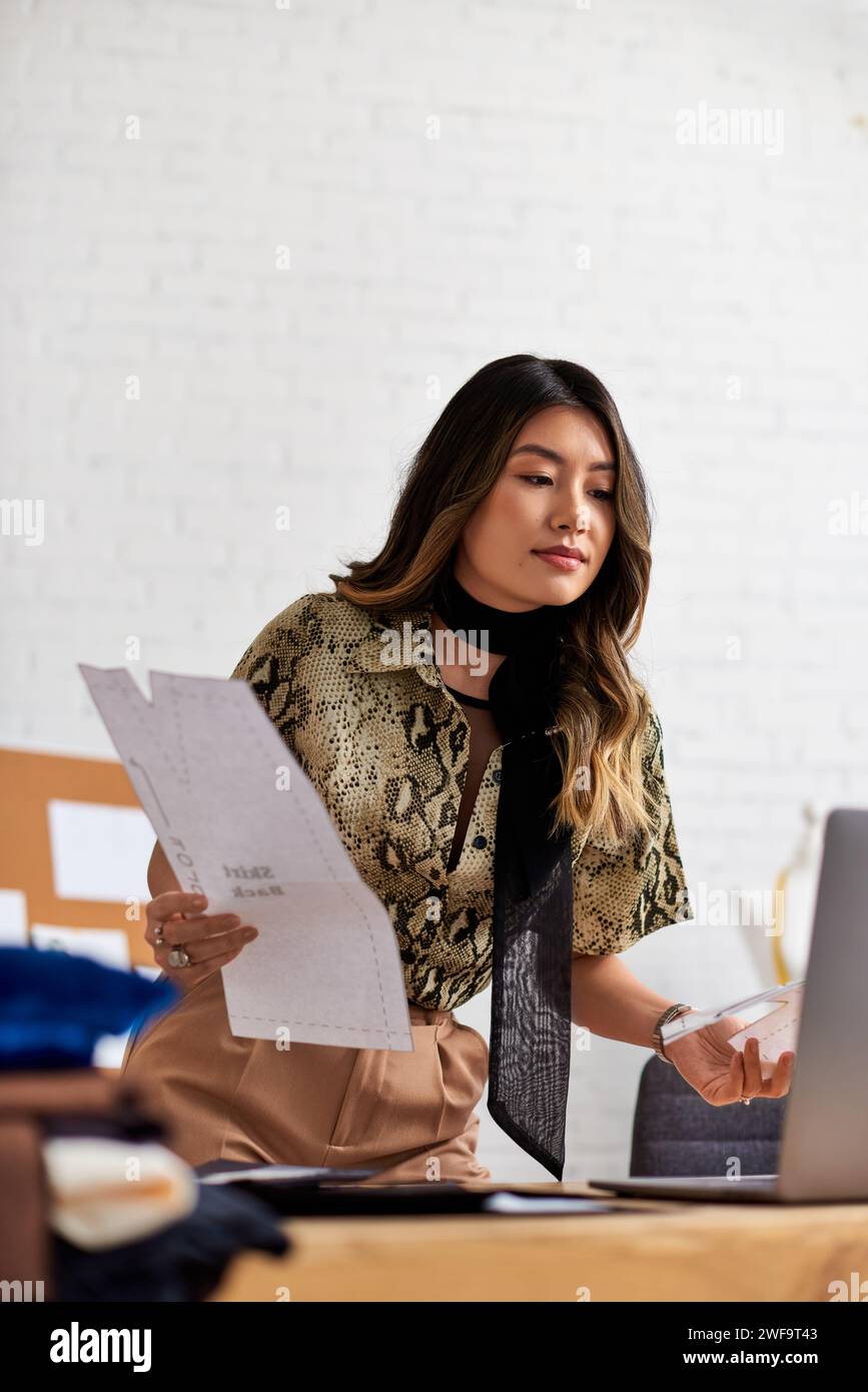 talentueux designer asiatique avec des patrons de couture regardant un ordinateur portable dans un atelier privé, une petite entreprise Banque D'Images