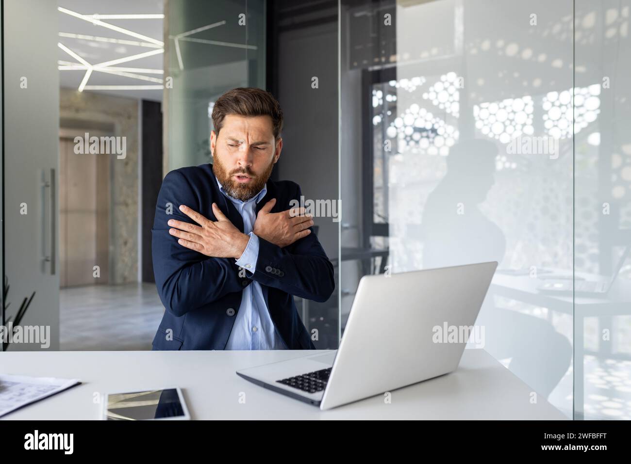 Inquiet jeune homme assis dans le bureau au bureau avec un ordinateur portable dans un costume et gelant, frissonnant du froid, s'étreignant et se réchauffant avec ses mains. Banque D'Images