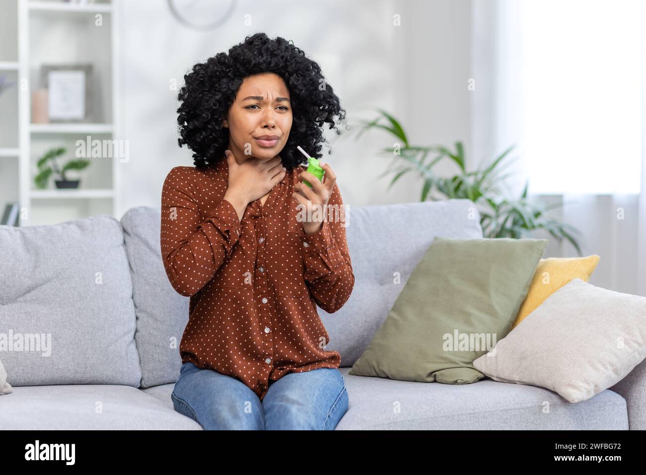 Jeune femme afro-américaine assise sur un canapé à la maison, tenant sa gorge, souffrant de douleurs et utilisant un spray médical. Banque D'Images