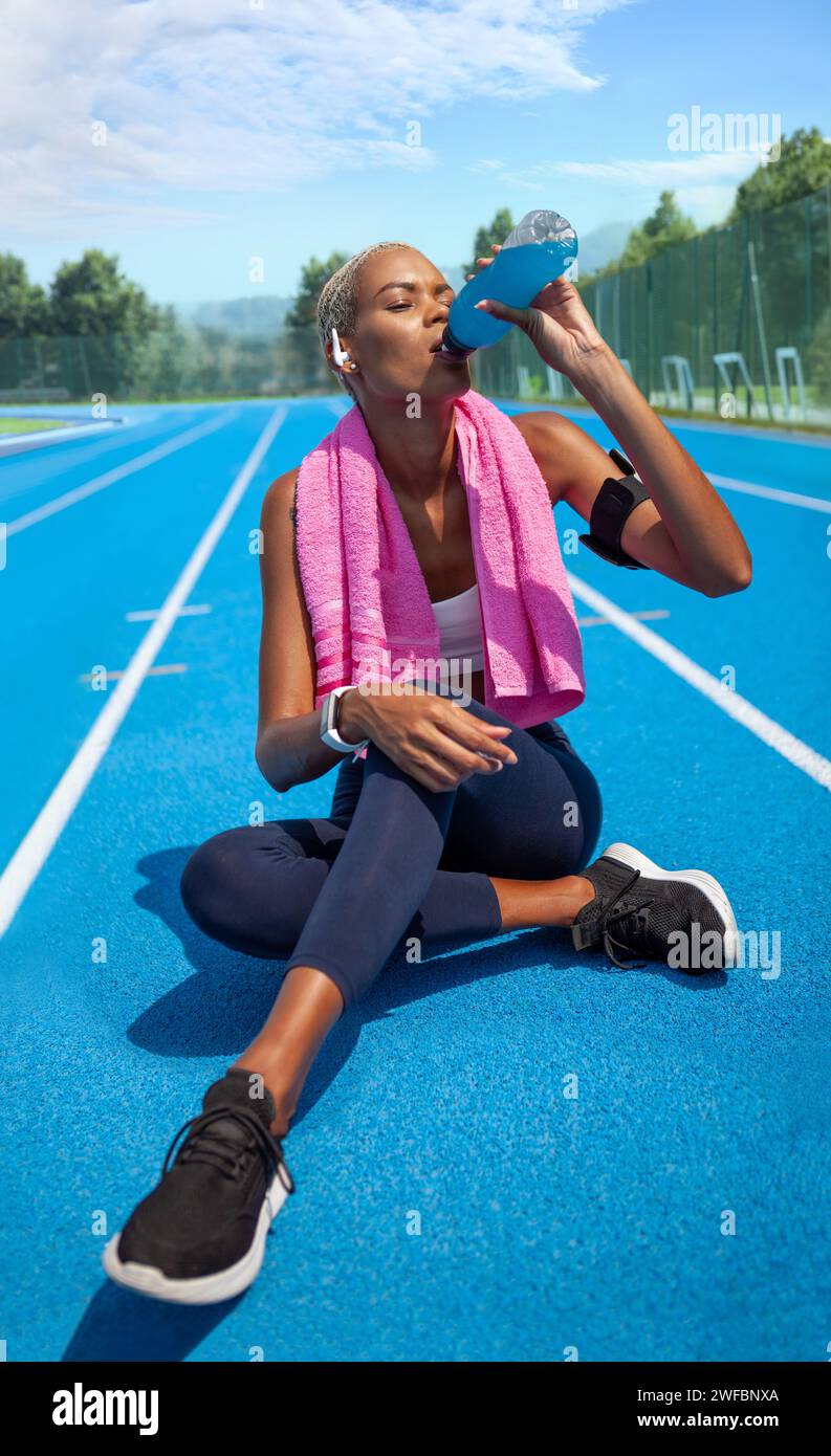 Athlète féminine souriante buvant une boisson énergétique à partir d'une bouteille après avoir couru un exercice sportif, porte des écouteurs, un brassard de smartphone et une serviette. Afro-américain Banque D'Images