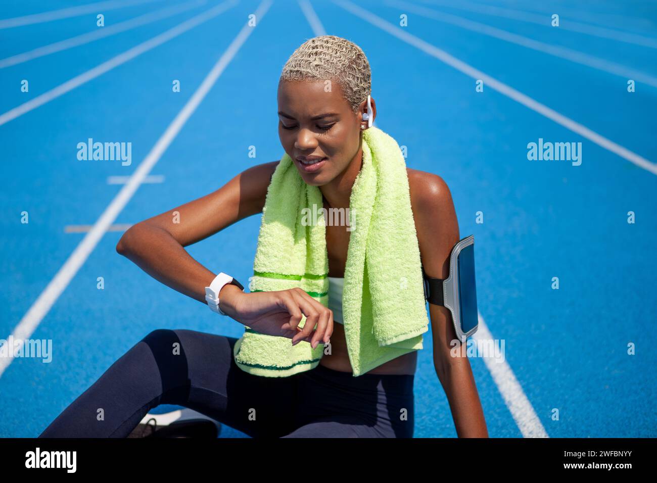 Athlète féminine souriante regarder une montre intelligente après l'exercice de sport de course, porte des écouteurs, un brassard de smartphone et une serviette. Femme afro-américaine assise Banque D'Images