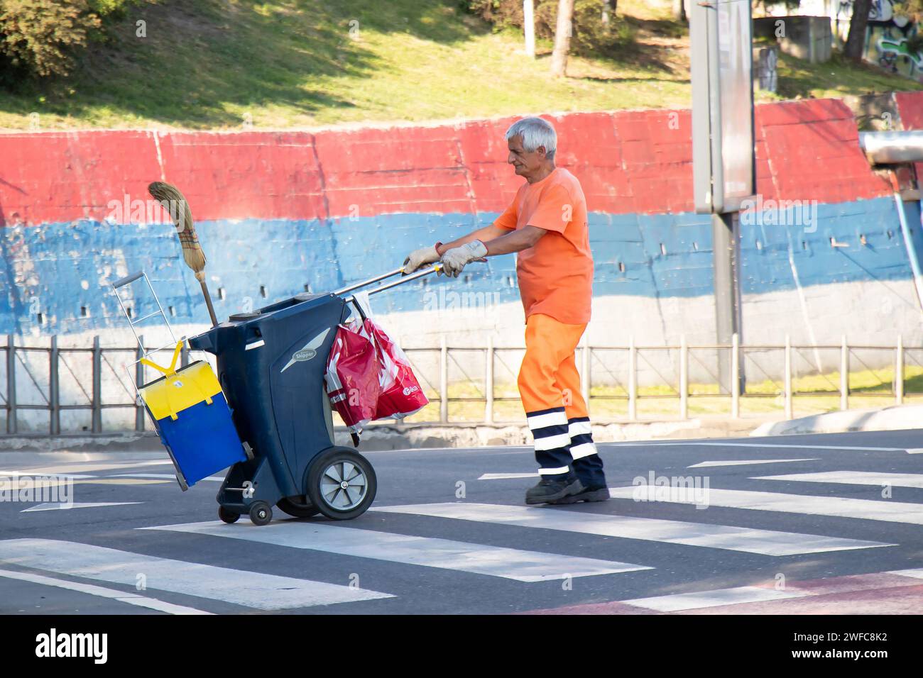Belgrade, Serbie - 5 juin 2021 : le collecteur d'ordures du service public de la ville traverse la rue tout en poussant le chariot de poubelle de taille avec balai et dustbi Banque D'Images