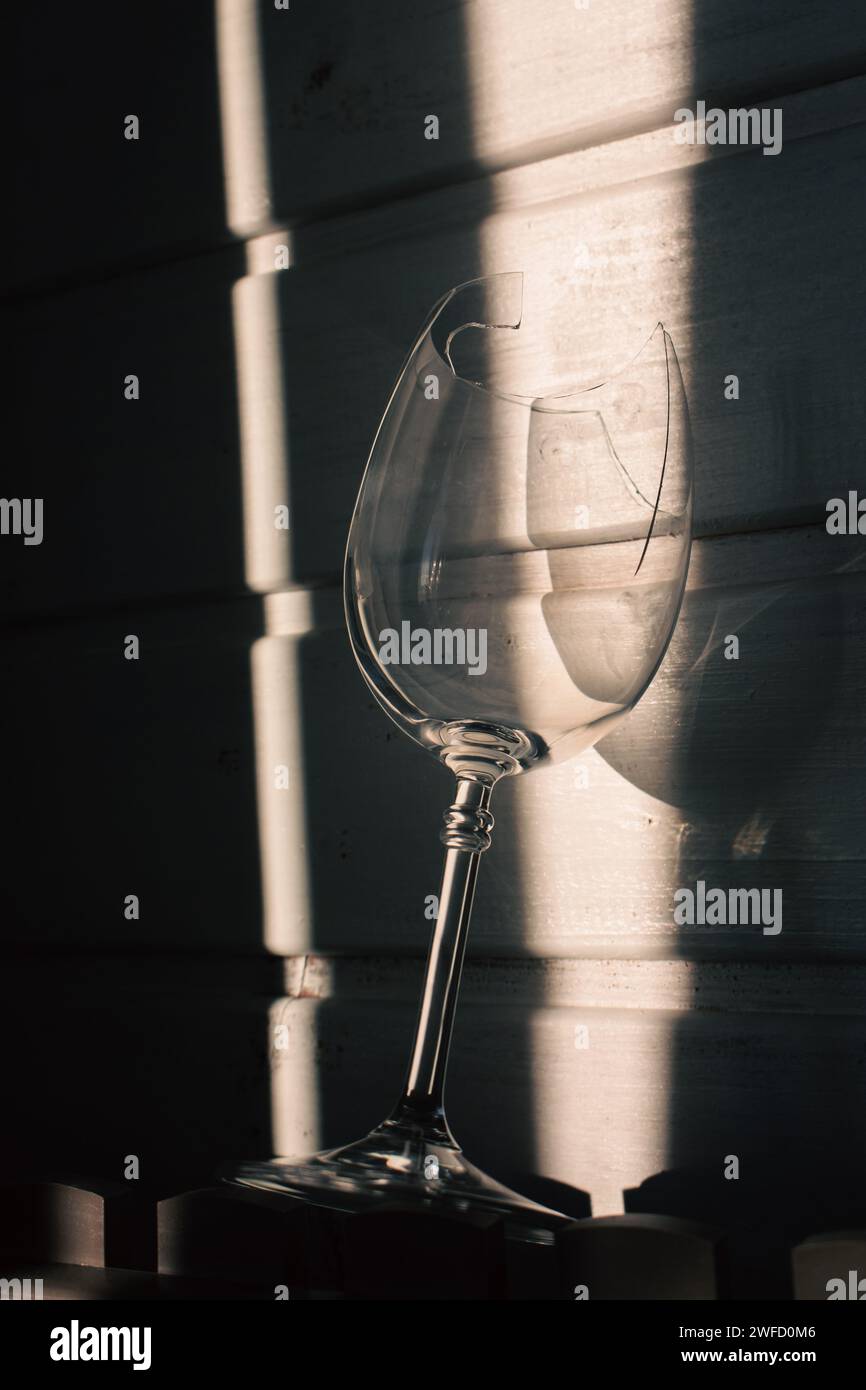 Verre à vin cassé dans la lumière du soleil sur fond en bois. Verre à vin endommagé avec bords tranchants. Verre fissuré dans les ombres. Nature morte créative. Objet fragile Banque D'Images