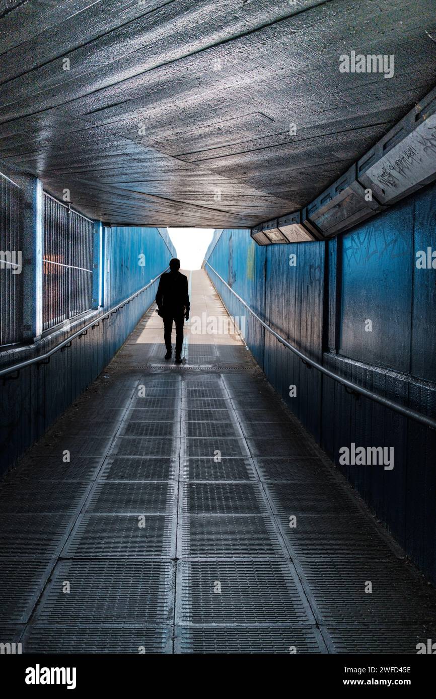 Personne en silhouette, marchant vers une lumière vive au bout d'un tunnel sombre avec des murs et un plafond métalliques, des graffitis et des grilles métalliques sur le sol. Banque D'Images