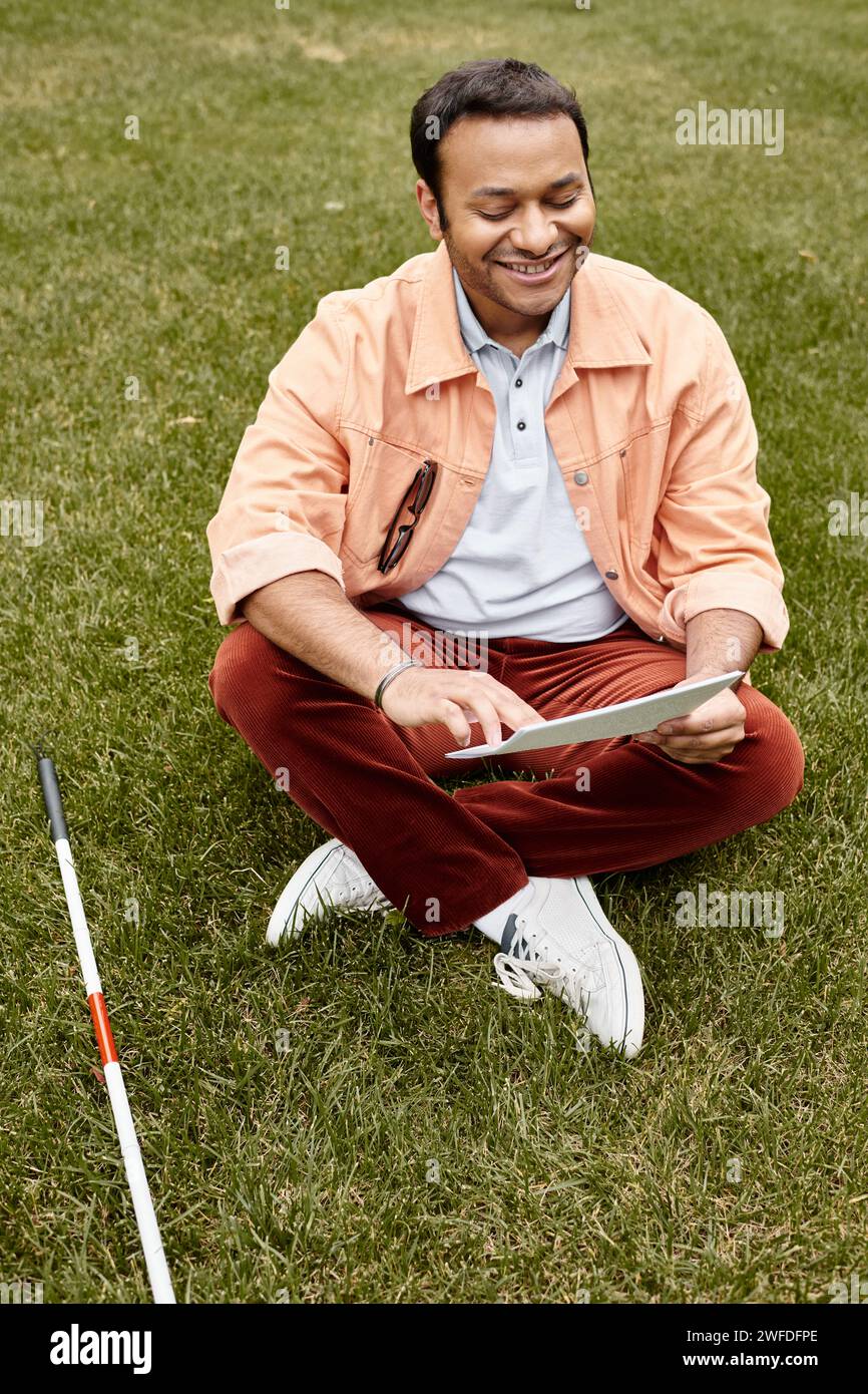 homme indien joyeux avec la cécité dans la veste orange assis sur l'herbe et lisant le code braille Banque D'Images