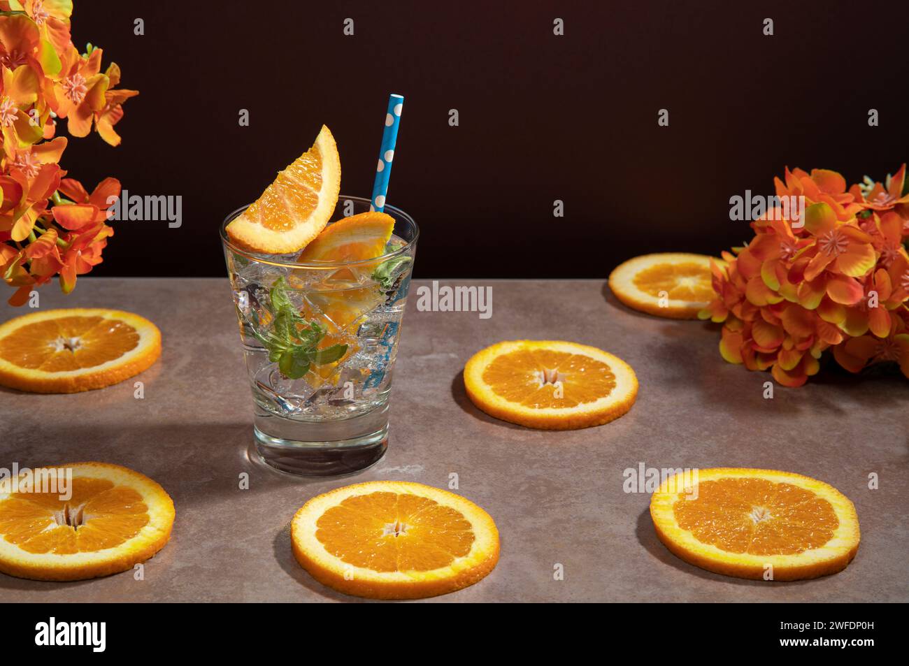 Cocktail en verre rempli de glace et de feuilles de menthe garnies de tranche d'orange et sorbet sur une table en céramique avec tranches d'orange contre rouge foncé Banque D'Images