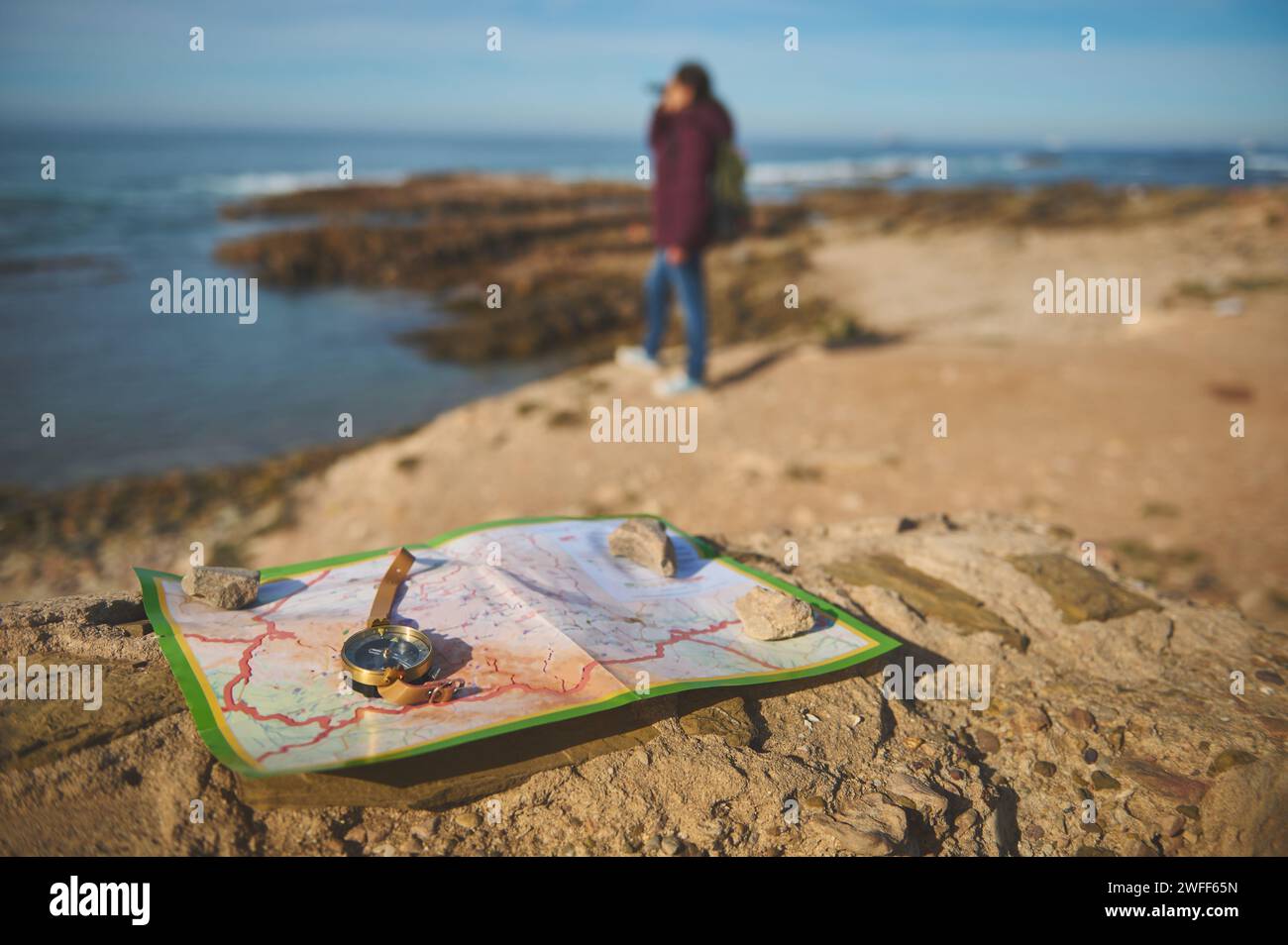 Détails sur une boussole et carte sur un rocher contre touriste féminine floue regardant dans la distance à travers des jumelles, debout sur le sommet d'une falaise, annonce Banque D'Images