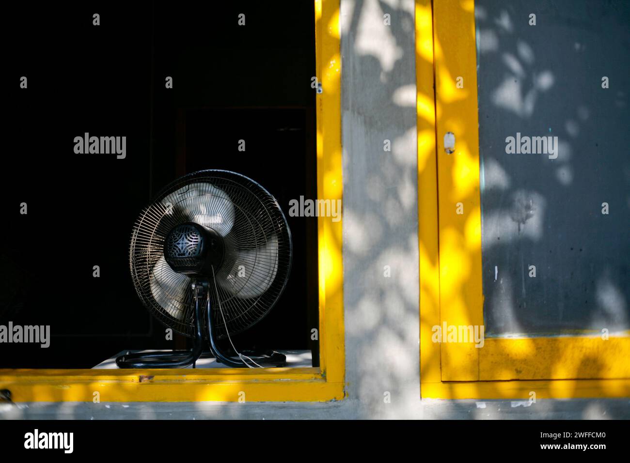 Ventilateur électrique noir dans la fenêtre jaune avec ombre de feuilles dans la journée d'été Banque D'Images