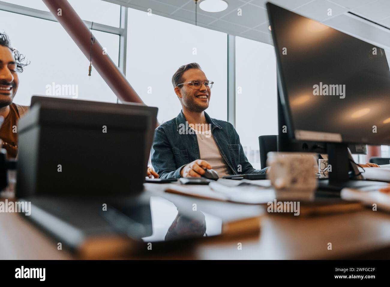 Sourire jeune homme d'affaires utilisant l'ordinateur tout en étant assis par collègue masculin travaillant au bureau dans le bureau Banque D'Images