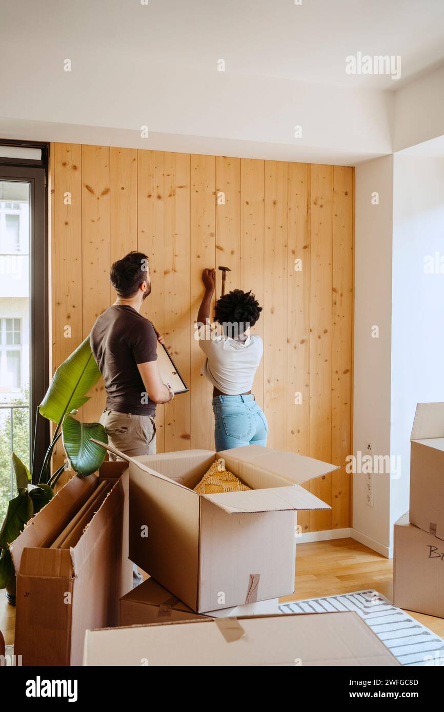 Femme martelant clou sur le mur en bois tout en se tenant avec petit ami à la nouvelle maison Banque D'Images
