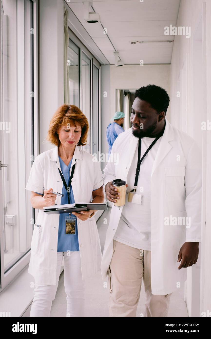 Médecin senior discutant sur le dossier médical avec un jeune collègue tout en marchant dans le couloir de l'hôpital Banque D'Images Médecin senior discutant sur le dossier médical avec un jeune collègue tout en marchant dans le couloir de l'hôpital Banque D'Images