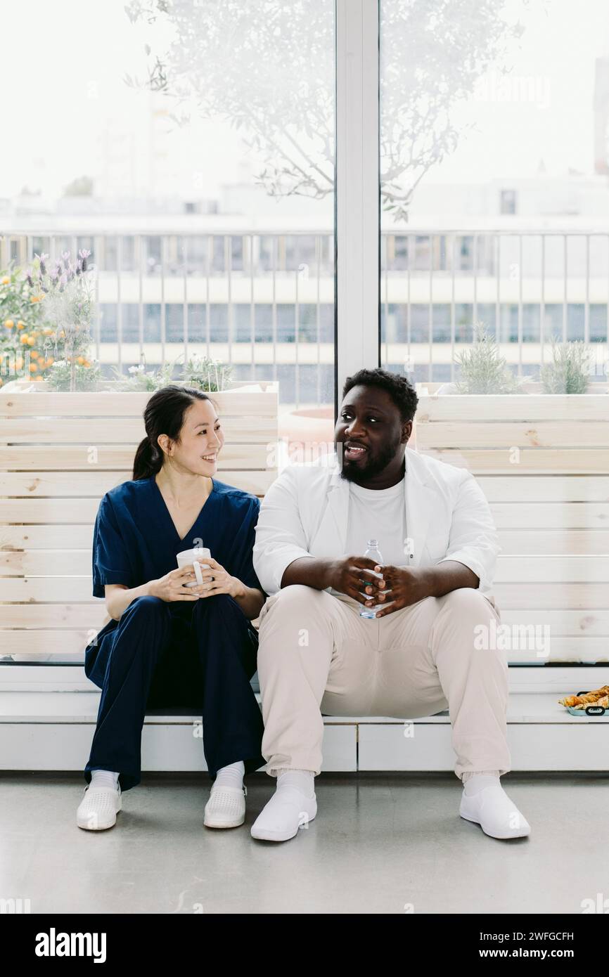 Longueur complète de collègues médicaux souriants hommes et femmes assis contre la fenêtre à l'hôpital Banque D'Images