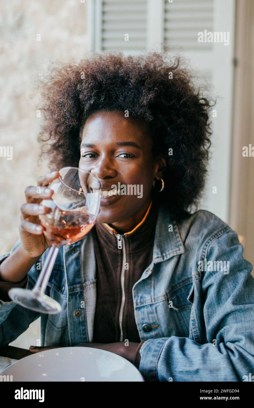 Portrait de jeune femme souriante avec coiffure afro buvant du vin au patio Banque D'Images