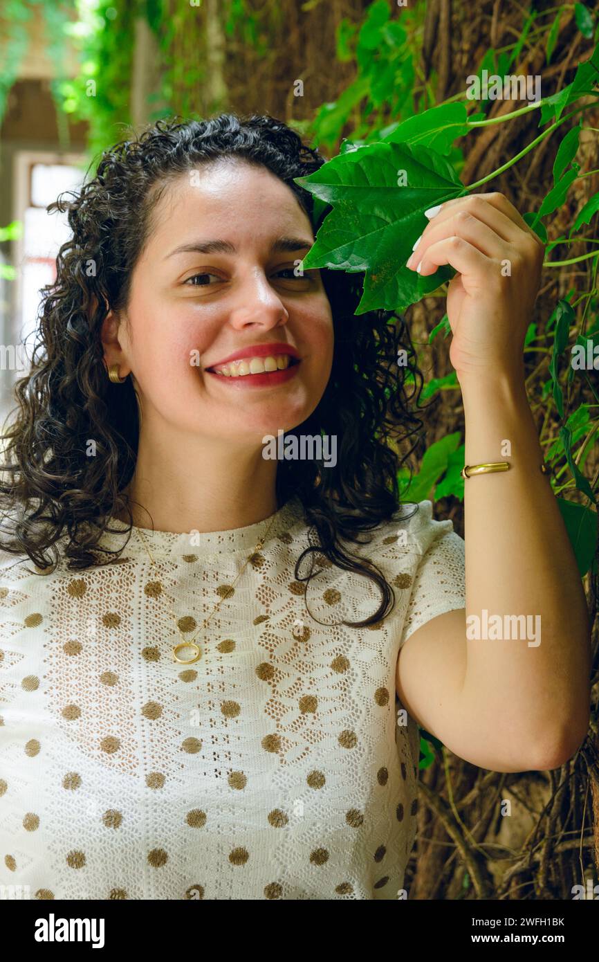 Portrait vertical de la jeune Latina vénézuélienne avec des bigoudis, heureux debout posant souriant regardant la caméra avec la feuille de plante grimpante sur le mur, la vie est Banque D'Images