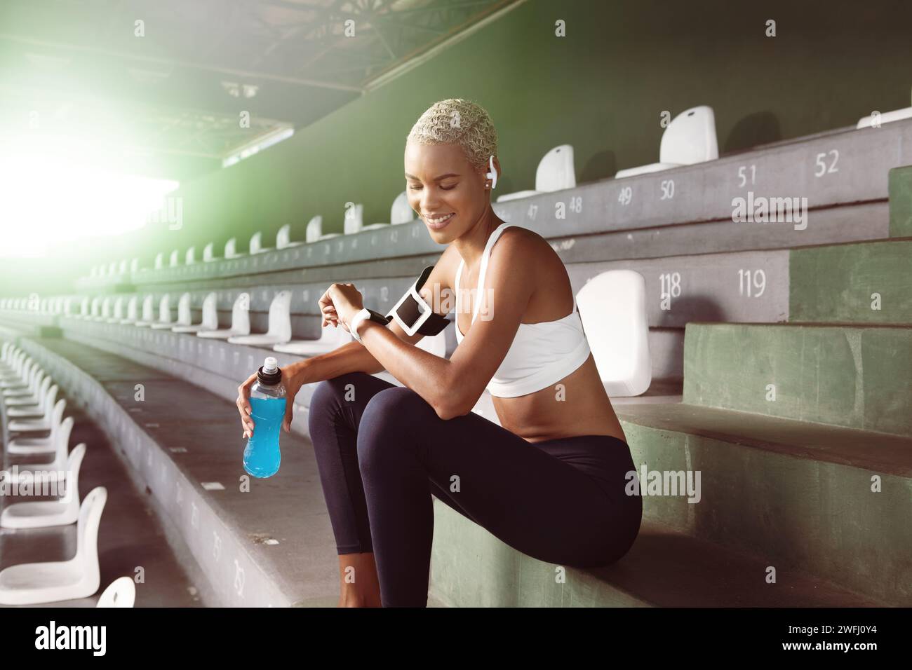 Athlète féminine souriante buvant une boisson énergétique de la bouteille après avoir couru l'exercice sportif, regardez smartwatch, porte des écouteurs. Femme afro-américaine Utiliz Banque D'Images