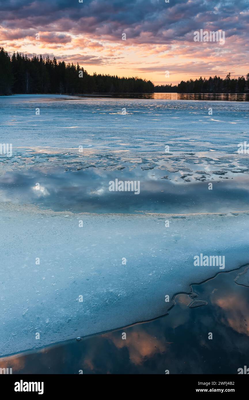 Une scène tranquille capture la beauté d'un lac partiellement gelé au crépuscule, avec des teintes subtiles du coucher de soleil se reflétant sur la glace et l'eau au milieu des s. Banque D'Images