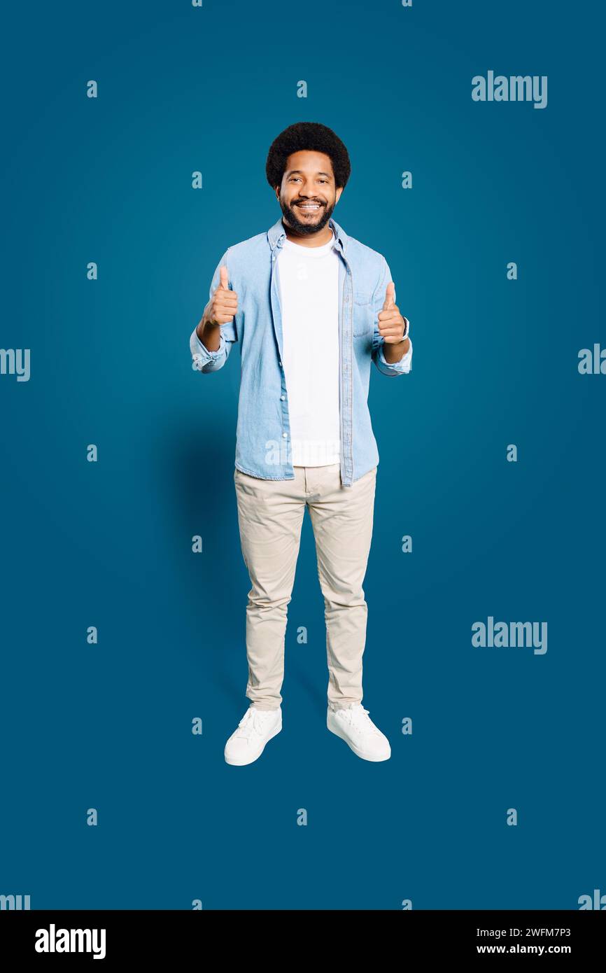 Un homme joyeux aux cheveux afro mousseux se tient sur toute la longueur, donnant deux pouces enthousiastes sur un fond bleu vibrant, symbolisant la positivité et l'approbation, Like-Sign, recommander Banque D'Images