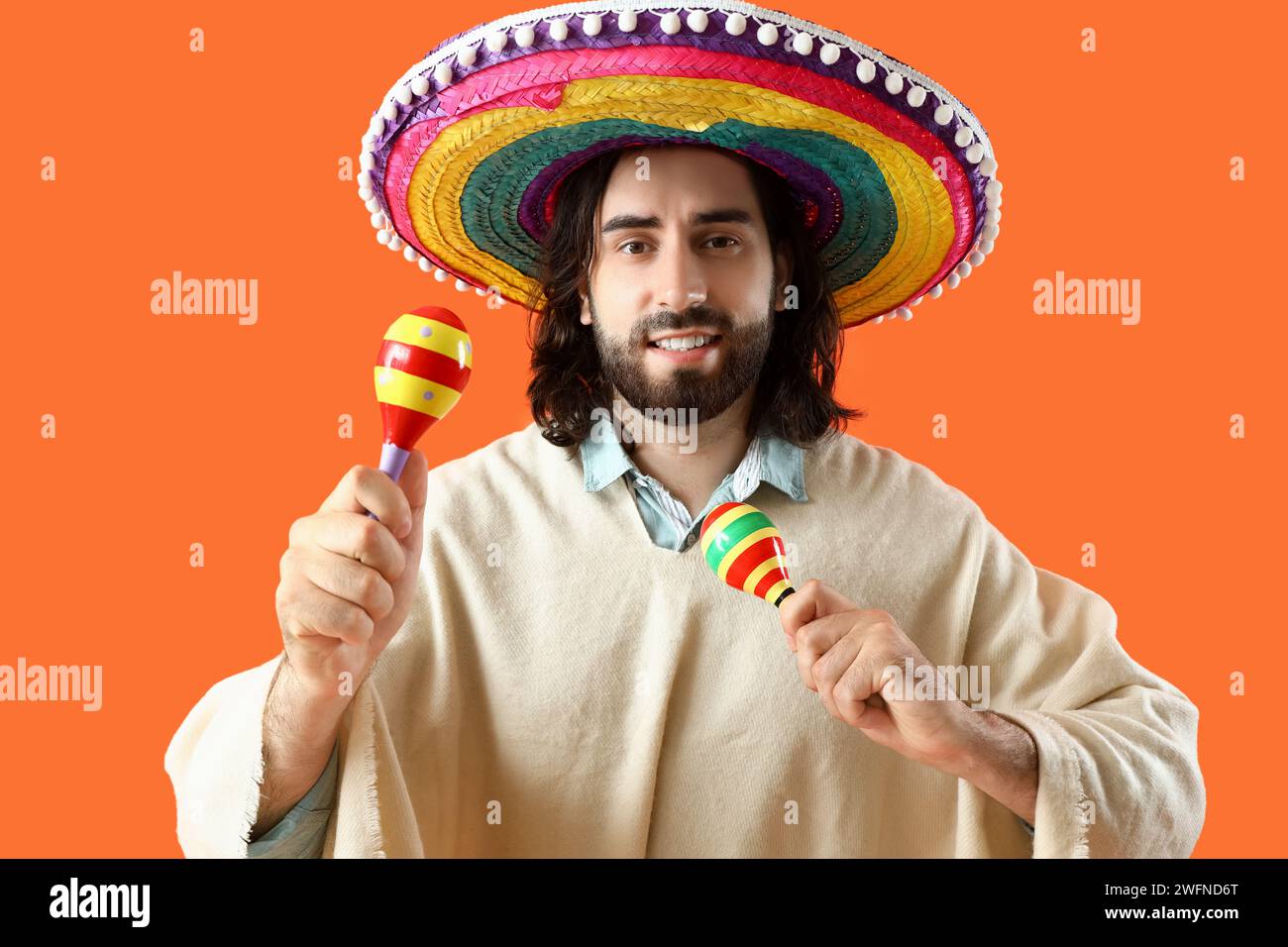 Beau jeune homme mexicain en sombrero et poncho jouant des maracas sur fond orange Banque D'Images