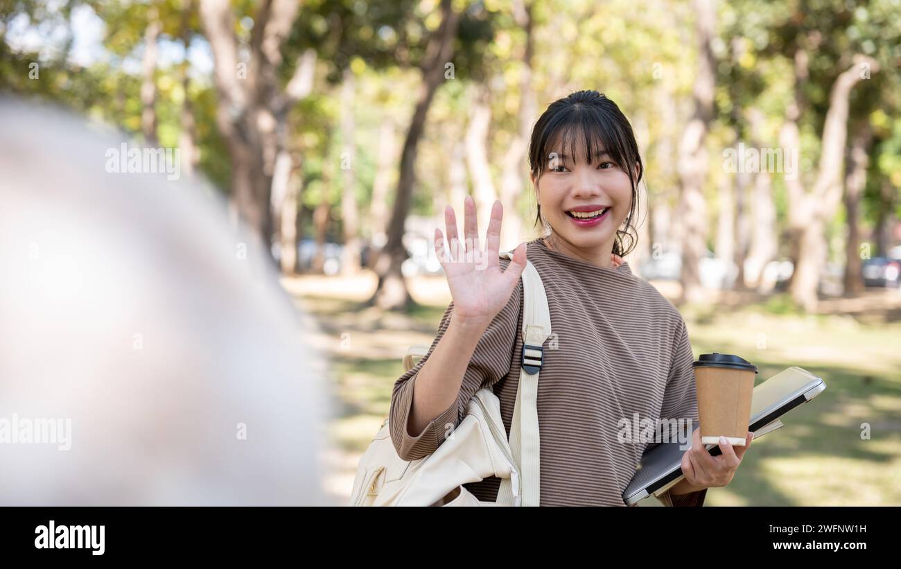 Une jeune étudiante asiatique amicale et mignonne agite la main, salue ou dit au revoir à son amie dans un parc. vie universitaire, école Banque D'Images