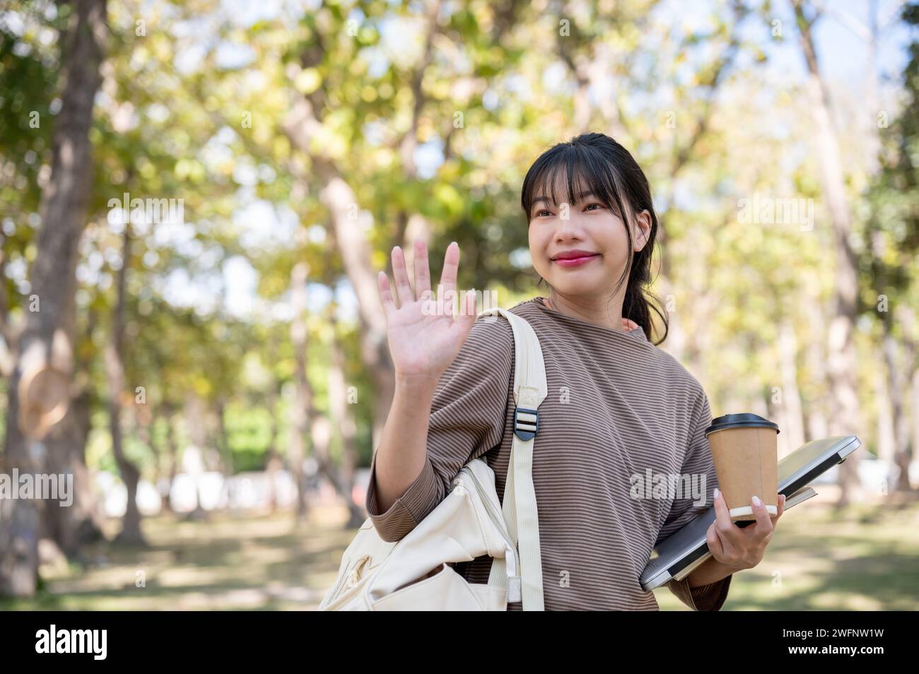 Une jeune étudiante asiatique amicale et mignonne agite la main, salue ou dit au revoir à son amie dans un parc. vie universitaire, école Banque D'Images