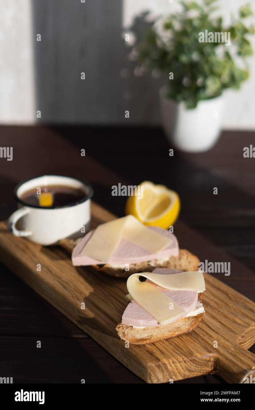 Sandwich avec beurre de jambon tranché et fromage sur planche à découper en bois près de tasse de thé avec citron Banque D'Images