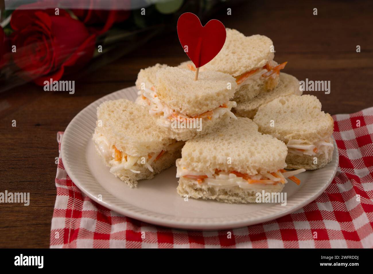 Sandwichs en forme de coeur faits de salade assis sur un plat blanc sur un tissu à carreaux rouge et blanc. Il y a un bouquet de roses sur le fond. saint-va Banque D'Images