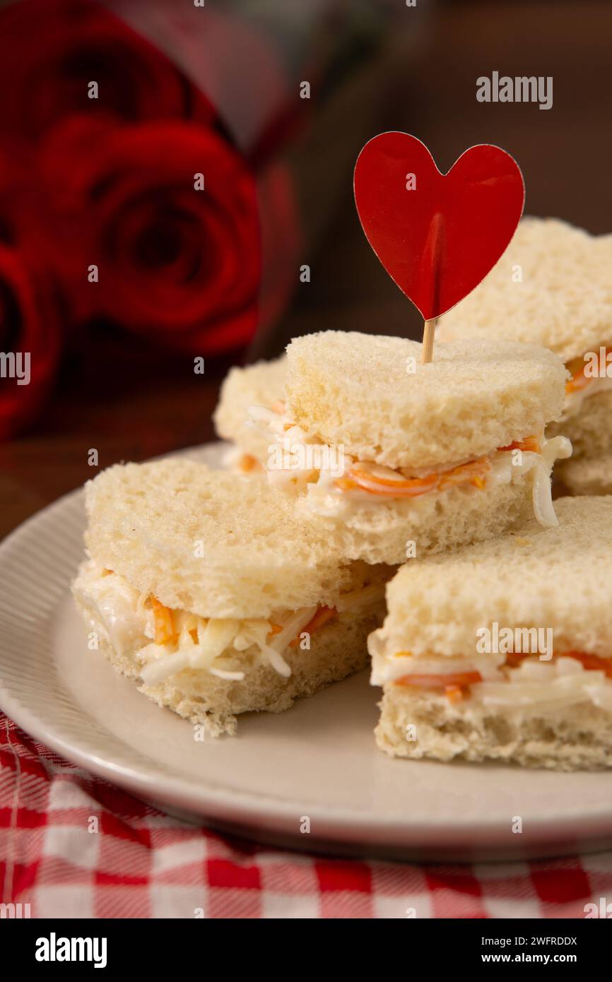Sandwichs en forme de coeur faits de salade assis sur un plat blanc sur un tissu à carreaux rouge et blanc. Il y a un bouquet de roses sur le fond. saint-va Banque D'Images