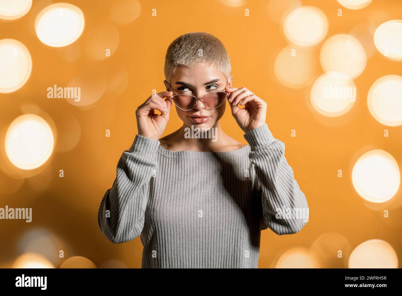 Jeune femme blonde avec shorthair regardant loin au coin tout en se tenant debout dans le pull à manches complètes avec ombre dans la lumière et l'ajustement des lunettes de soleil sur le nez Banque D'Images