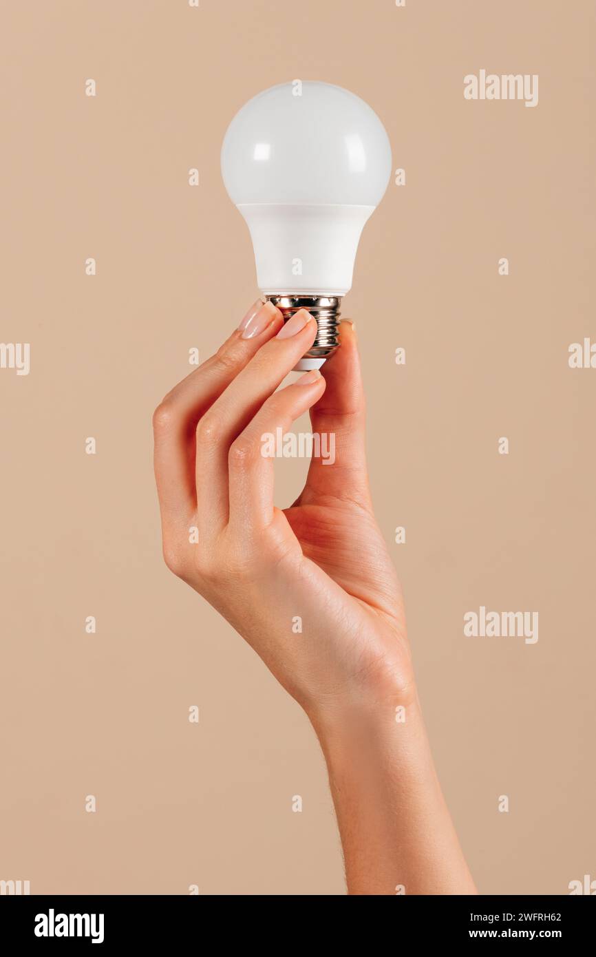 Main de jeune femme anonyme montrant ampoule LED blanche dans le concept d'écologie sur fond beige en studio Banque D'Images