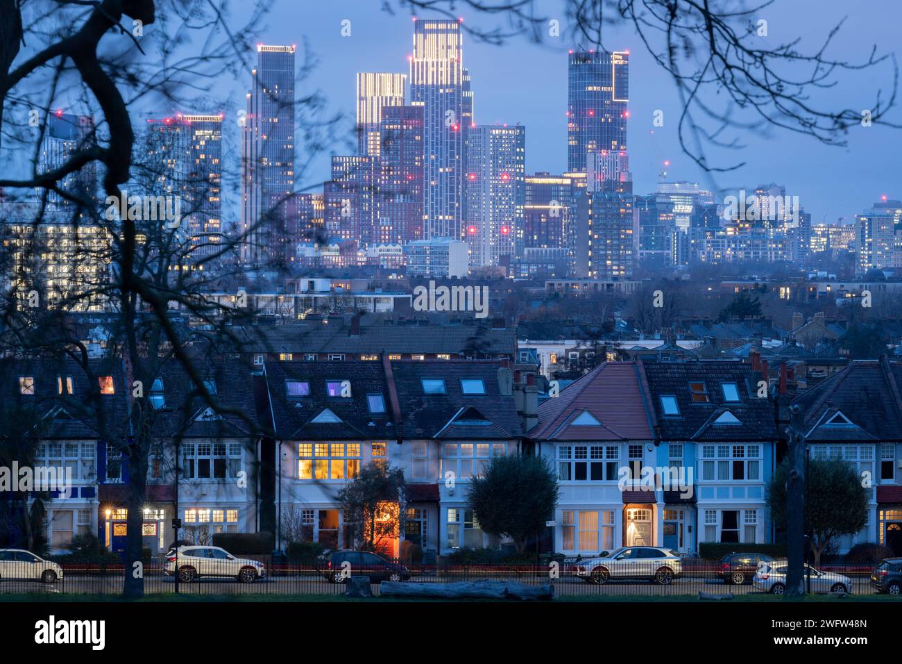 Vu à travers les branches des arbres dans Ruskin Park sont les porches éclairés des maisons d'époque en terrasses et au loin, le développement croissant à Nine Elms, le 31 janvier 2024, à Londres, en Angleterre. Banque D'Images