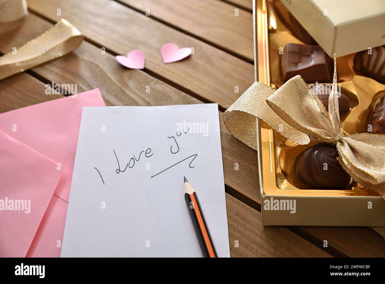 Note Je t'aime écrit au crayon sur une table en bois avec boîte de chocolats et découpes de coeurs roses. Vue de dessus surélevée. Banque D'Images