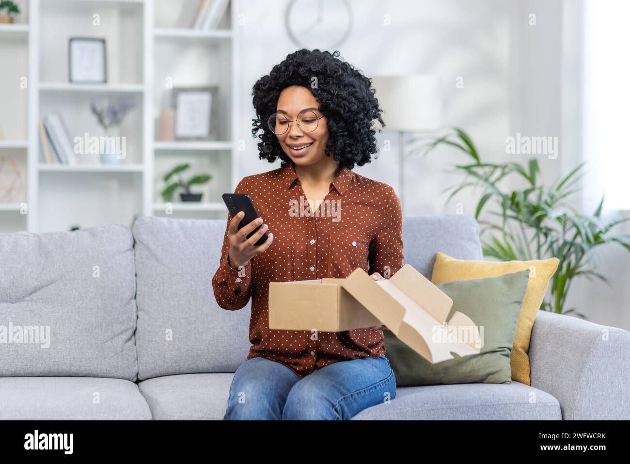 Heureuse jeune femme afro-américaine assise sur un canapé à la maison, tenant la boîte de colis de livraison ouverte et en utilisant le téléphone portable souriant. Banque D'Images