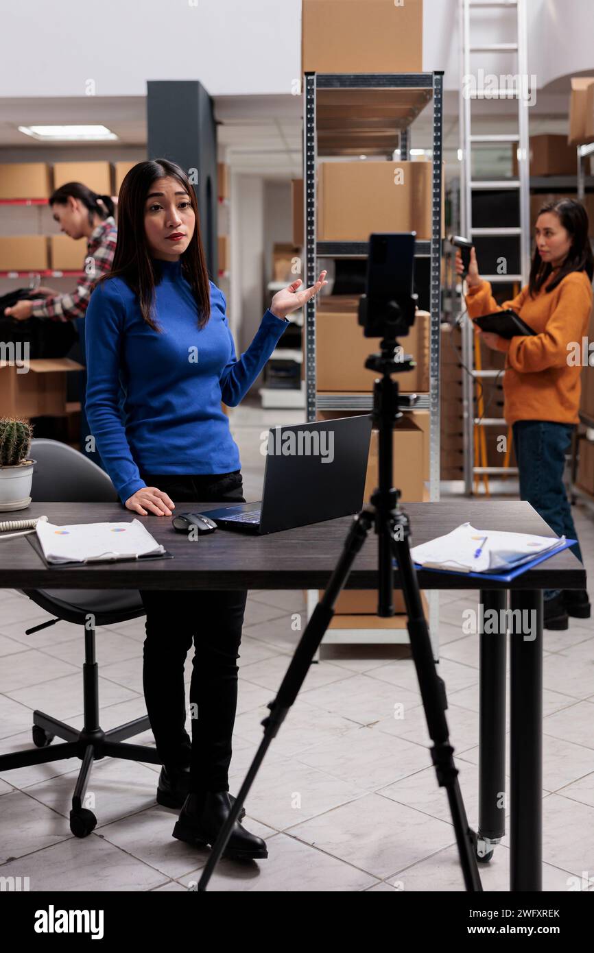 Entrepôt jeune femme asiatique Manager enregistrant une vidéo de marketing pour promouvoir le produit. Un employé de magasin filme l'équipe de préparateurs de commandes préparant le colis pour l'expédition sur smartphone Banque D'Images