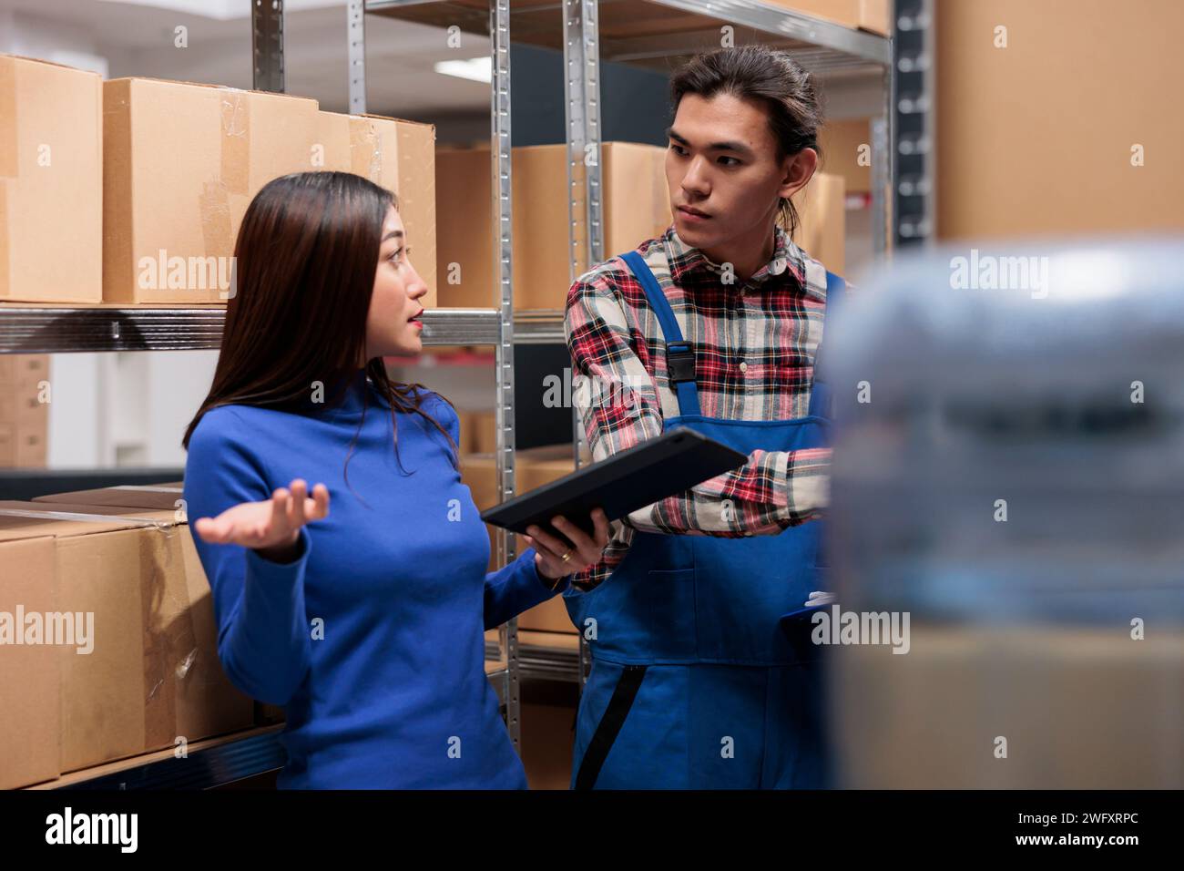 Les employés asiatiques de l'entrepôt discutent du suivi des stocks tout en gérant l'inventaire des produits dans la salle de stockage. Les employés de Storehouse font la gestion financière sur tablette numérique et parlent près de l'étagère des boîtes Banque D'Images