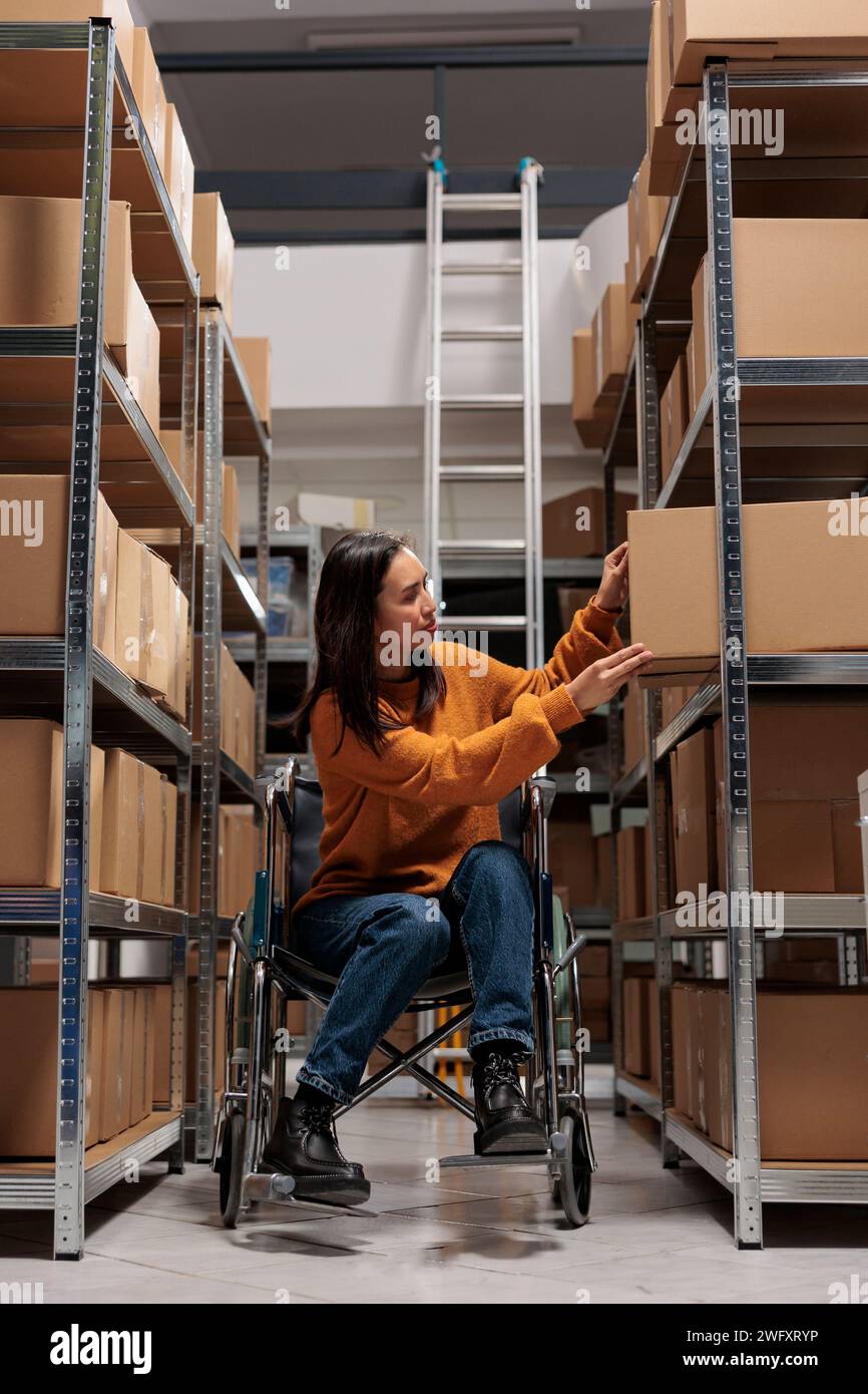 Magasin préparateur de commande asiatique en fauteuil roulant prenant le colis du rack pour emballer le produit avant expédition. Magasin d'affaires de détail jeune femme gestionnaire de paquet choisissant la boîte sur l'étagère Banque D'Images