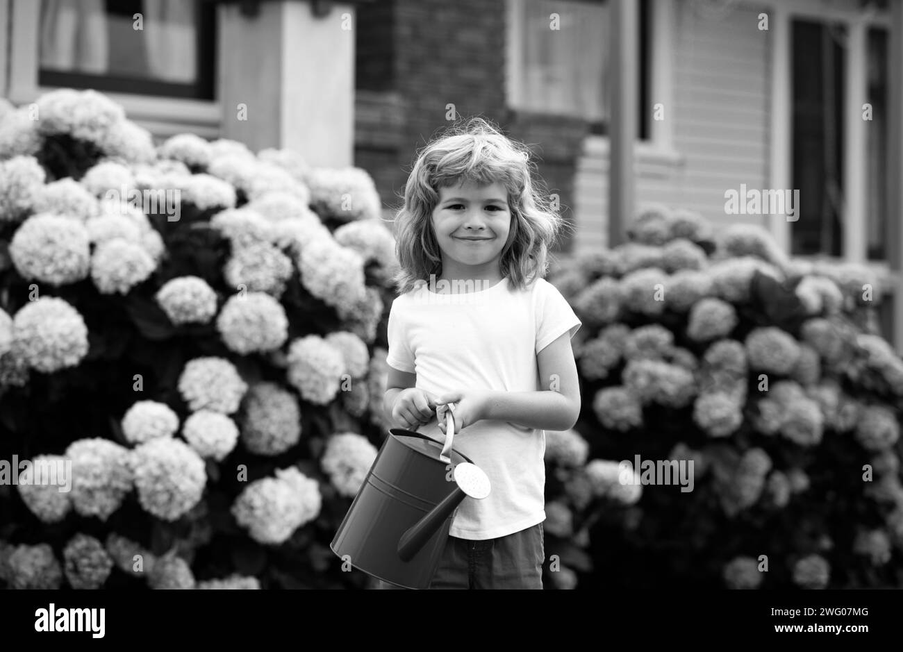 Joli petit garçon arrosoir fleurs avec arrosoir dans le jardin. Enfant habillé en été clair ferme et t-shirt coloré, souriant et s'amuser. Banque D'Images